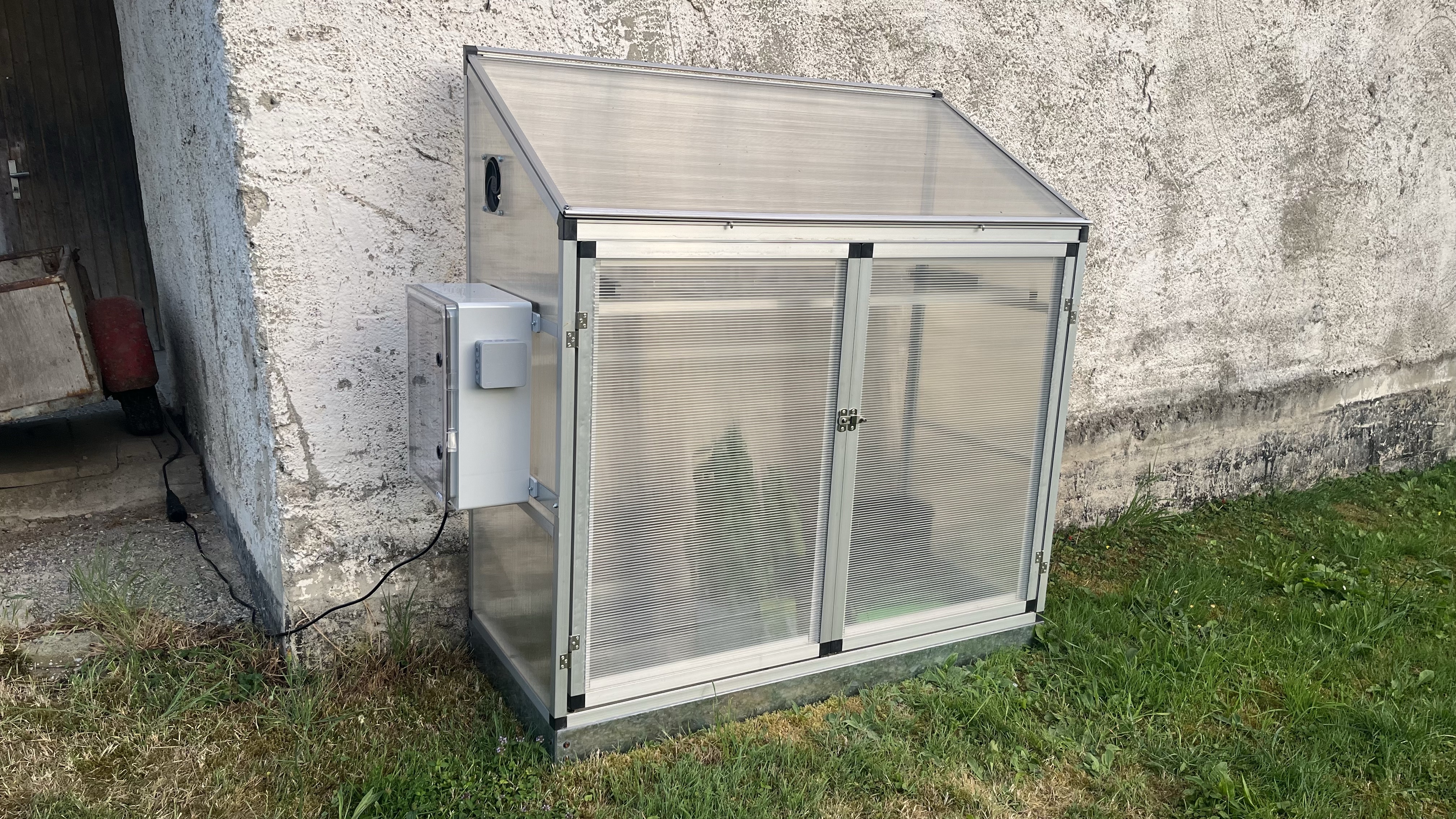 Ein Gewächshaus mit Schaltkasten und Technik zu Automatisierung von Temperatur und Bodenfeuchtigkeit